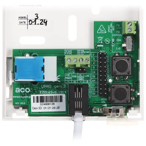 Zdjęcie przedstawia zieloną płytkę drukowaną z różnymi komponentami elektronicznymi, w tym złączami, przyciskami i oznaczeniami technicznymi. Płytka jest zamontowana w białej obudowie z widocznymi oznaczeniami montażowymi i datą.