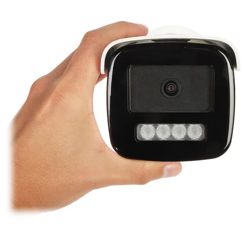 Osoba trzyma w ręku czarną kamerę monitoringu z białą obudową, która ma widoczne diody LED poniżej obiektywu. Kamera jest skierowana w stronę obiektywu aparatu, ukazując szczegóły jej przedniej części.