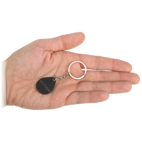Czarny brelok z numerem seryjnym, przymocowany do metalowego kółka na klucze, leży na otwartej dłoni. Produkt jest kompaktowy i łatwy do przenoszenia.