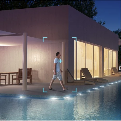 Na zdjęciu widać nowoczesny budynek z dużymi oknami, oświetlony w nocy, z basenem na pierwszym planie. Mężczyzna w jasnym ubraniu przechodzi obok basenu, a na zdjęciu zaznaczono niebieski prostokąt sugerujący funkcję śledzenia kamery.