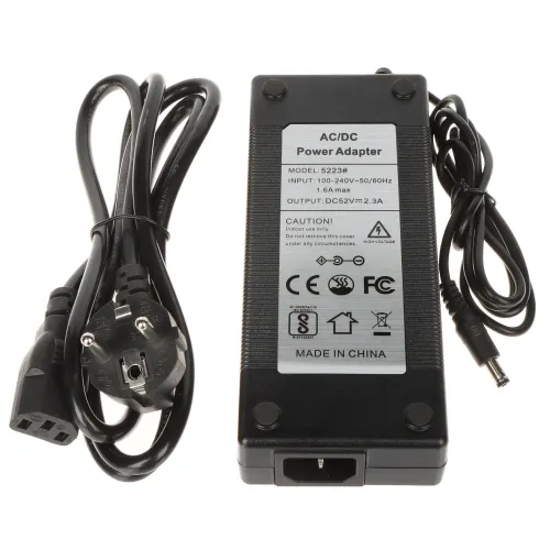 Na zdjęciu znajduje się czarny zasilacz AC/DC z etykietą informacyjną na górze, obok którego leżą dwa przewody z wtyczkami. Etykieta zawiera specyfikacje techniczne, takie jak model, napięcie wejściowe i wyjściowe oraz certyfikaty bezpieczeństwa.