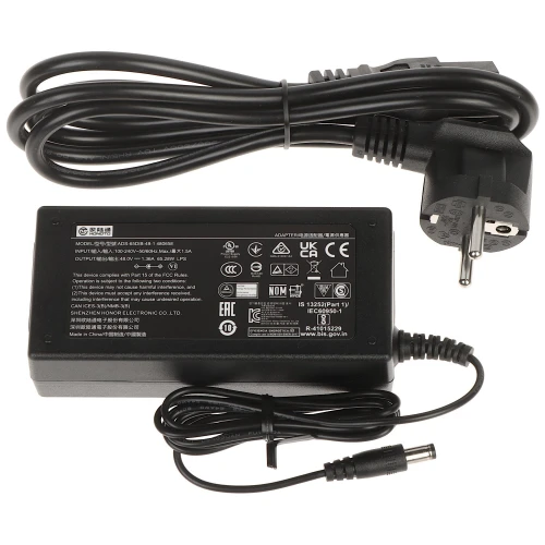 Na zdjęciu znajduje się czarny zasilacz sieciowy z etykietą techniczną, obok którego leży odłączany kabel zasilający z wtyczką. Zasilacz ma prostokątny kształt i wyposażony jest w przewód z okrągłą końcówką do podłączenia urządzeń.