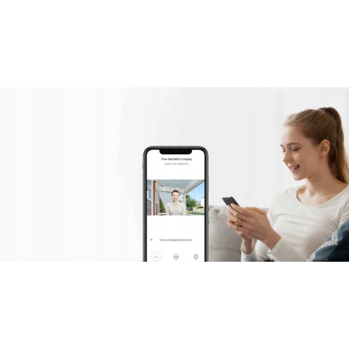 Na zdjęciu widać kobietę siedzącą na kanapie, trzymającą smartfon, na którym wyświetlany jest obraz z elektronicznego wizjera drzwiowego. Ekran telefonu pokazuje osobę stojącą przed drzwiami, co sugeruje funkcję podglądu w czasie rzeczywistym.