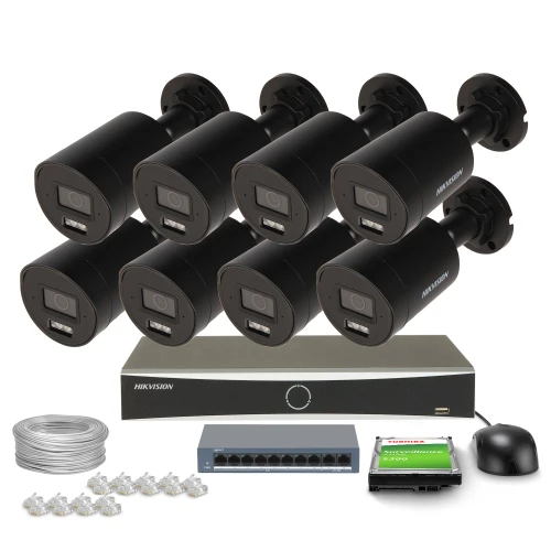 Na zdjęciu znajduje się zestaw do monitoringu składający się z ośmiu czarnych kamer, rejestratora, kabla, przełącznika sieciowego, myszy komputerowej oraz karty pamięci. Kamery są ułożone w dwóch rzędach, a pozostałe elementy zestawu są rozmieszczone poniżej.
