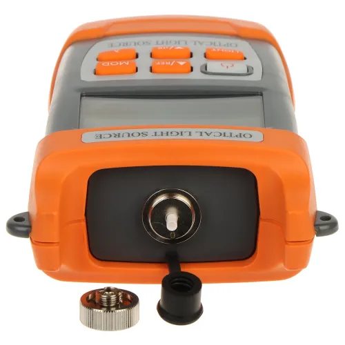 Na zdjęciu znajduje się pomarańczowo-szare urządzenie z wyświetlaczem i przyciskami, oznaczone jako "OPTICAL LIGHT SOURCE". Przed urządzeniem leżą dwa małe elementy, prawdopodobnie części złącza optycznego.